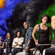 The cast of F9 lean on cars with colorful smoke behind them.